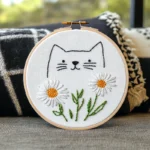 A close-up of an artistic embroidery piece featuring a cat and daisies