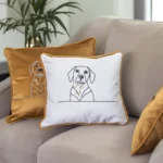 A gold and a white throw pillow on a khaki-colored sofa, with the white pillow featuring an abstract art dog embroidery.