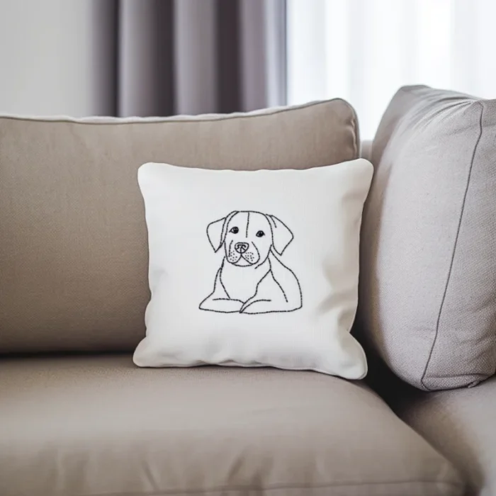 A white throw pillow on a khaki-colored sofa, featuring an embroidered dog gazing into the distance.