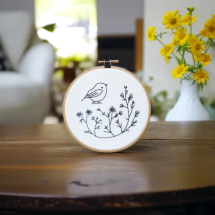 A bird and branch embroidery piece placed on a table, with a sofa and sunflowers in the background