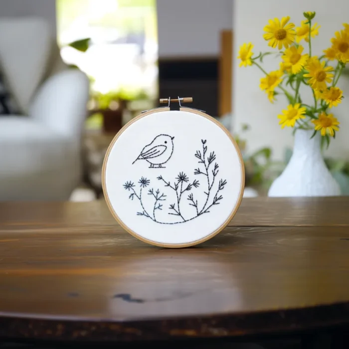 A bird and branch embroidery piece placed on a table, with a sofa and sunflowers in the background