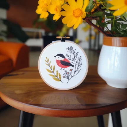 A bird and tree embroidery piece placed on a table alongside a pot of daisies