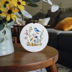 A coffee table with a blue bird and floral embroidery piece alongside a small vase of sunflowers