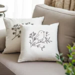 Two white fabric pillows with black line art bird embroidery placed on a light gray sofa