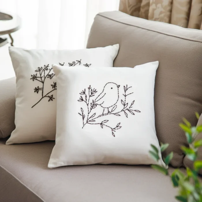 Two white fabric pillows with black line art bird embroidery placed on a light gray sofa