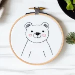 Adorable blushing bear embroidery in a hoop, stitched on clean white fabric in soft lighting