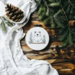 Bear and pine tree embroidery placed on a table, with white fabric and pine branches adding a cozy, rustic touch