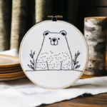 Vertical embroidery artwork of a small bear and wildflowers on a piece of white fabric