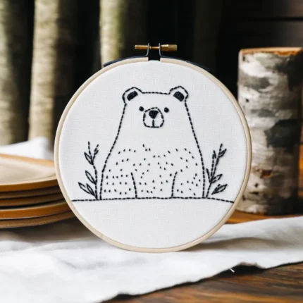 Vertical embroidery artwork of a small bear and wildflowers on a piece of white fabric