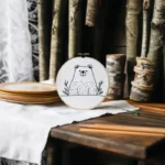 Table with hand bear embroidery artwork, white fabric, paint palette, brushes, sheer curtains, and wooden decor