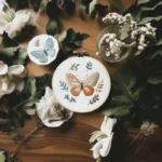table with handmade embroidery piece, white flowers, and green leaves