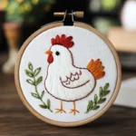 Round embroidery hoop on wooden table featuring a white chicken with green leaves