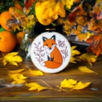 fox embroidery artwork placed on the ground with leaves and oranges