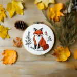 fox embroidery piece on the ground, decorated with autumn leaves and pine branches