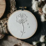 hand embroidery of a dandelion design in white thread placed on a table