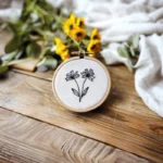 a wildflower embroidery displayed on a wooden table, with small yellow flowers beside it