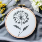 black and white dandelion line embroidery displayed on a blue-gray fabric