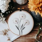 three dandelion flowers hand embroidered on white fabric held by an embroidery hoop