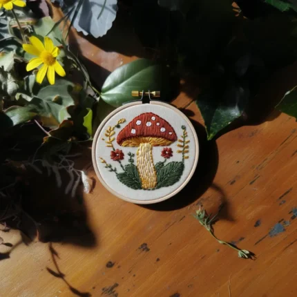 hand-stitched red mushroom embroidery hoop displayed on a wooden table surrounded by dark green plants and a yellow flower