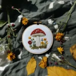red mushroom and floral embroidery in a hoop on dark green fabric with scattered autumn leaves