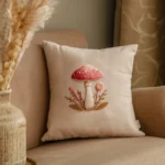 white decorative cushion featuring a stitched red mushroom and pink floral design resting on a cozy beige sofa
