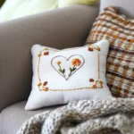 white decorative cushion featuring an orange floral heart embroidery and stitched border on a cozy grey sofa